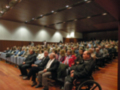 Una audiencia en un auditorio lleno de personas sentadas en filas, algunos en sillas y otros en sillas de ruedas. El ambiente es formal con iluminación adecuada para la sala.
