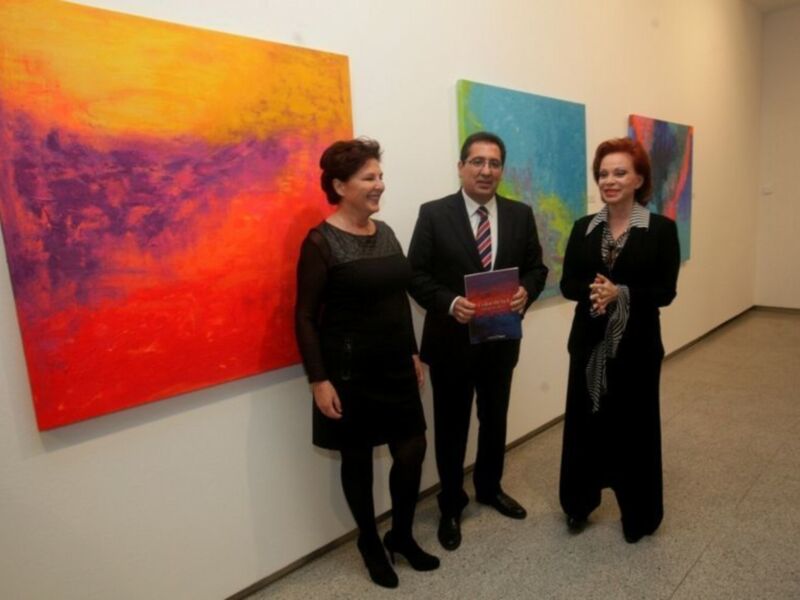 Tres personas posan frente a dos obras de arte abstractas vibrantes, una con colores rojos y naranjas, la otra con tonos azules y verdes. La persona central sostiene un libro.