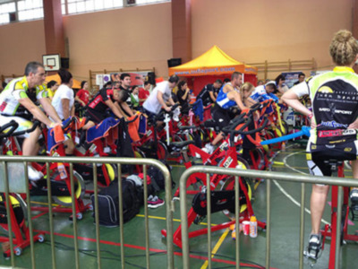 Una escena en un gimnasio de ciclismo indoor con atletas en bicicletas estáticas, un toldo amarillo y una canasta de baloncesto en el fondo.