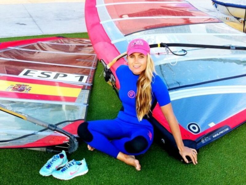 Una mujer en traje de neopreno azul y gorra roja posa junto a varios tablas de windsurf con la bandera española.