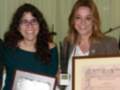 Dos mujeres sonríen mientras reciben un premio y un diploma.