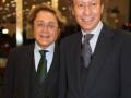 Dos hombres en trajes posan juntos, uno con una chaqueta de rayas y el otro con un chaleco. Ambos tienen camisas blancas y corbatas. El fondo es de noche con luces interiores.