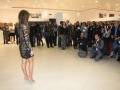 Una mujer con vestido negro y tacones altos se encuentra en el centro de una sala llena de personas. Algunas están tomando fotos con cámaras y teléfonos móviles, mientras otros observan la escena. La sala tiene una decoración moderna y una pared con fotos enmarcadas.