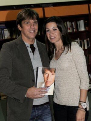 Un hombre y una mujer sonríen mientras sostienen un libro. El hombre lleva una camisa de manga larga y vaqueros, mientras que la mujer usa una blusa con estrellas y pantalones negros. La imagen está ambientada en un entorno de biblioteca o sala de lectura, con estanterías llenas de libros al fondo.