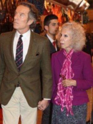 Una mujer con una chaqueta púrpura y un pañuelo rosa, caminando junto a un hombre con una chaqueta verde y corbata roja. Ambos están en un ambiente formal, posiblemente una gala o evento importante.