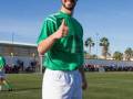 Un joven con una camiseta verde y pantalones cortos blancos, sonriendo y dándole un pulgar hacia arriba en una cancha de fútbol. El fondo muestra otros jugadores y palmeras bajo un cielo azul claro.
