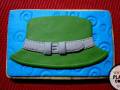 Un pastel decorado con forma de sombrero verde y cinturón blanco sobre un fondo azul.