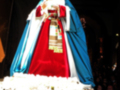 Imagen de una Virgen María vestida con trajes tradicionales, rodeada por flores y gente en el fondo.
