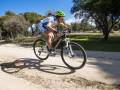 Un ciclista monta una bicicleta de montaña en un camino de tierra, rodeado por árboles y vegetación. El cielo está despejado con algunas nubes.