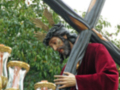 Un personaje vestido con un manto rojo sostiene una cruz, posiblemente en una procesión religiosa, con árboles verdes al fondo.