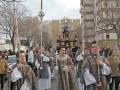 Imagen de una procesión religiosa con personas vestidas en trajes tradicionales, portando palos y una imagen sobre un carro. Fondo urbano con edificios de varios pisos.