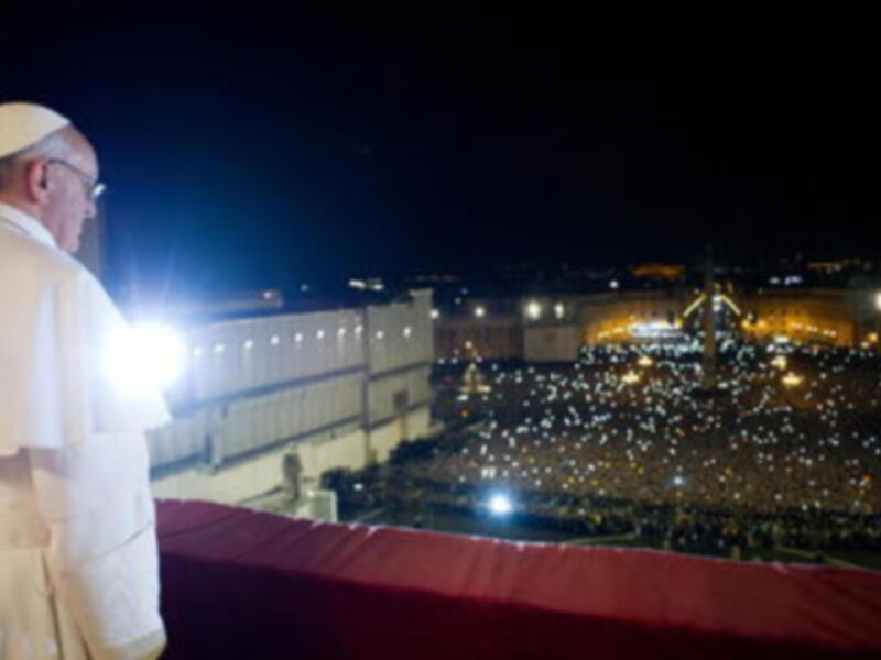 El Papa Francisco, vestido con su hábito bíblico, se encuentra en una plataforma elevada frente a un gran público iluminado por luces. La imagen captura el momento en que el Papa dirige su mensaje a los fieles reunidos, con la arquitectura y el cielo nocturno del Vaticano al fondo.
