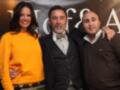 Tres personas posan juntas en una foto. La persona de la izquierda lleva un suéter naranja y pantalones con estampado. La persona en el centro lleva un traje oscuro y corbata de lazo. La persona de la derecha lleva un suéter marrón y camisa blanca. El fondo muestra letras que parecen "VIAFAR".