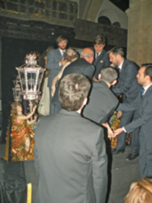 Un grupo de personas, algunos con trajes formales y otros en ropa casual, están rodeando una estatua de un santo. La estatua parece ser de madera y está colocada en una base con un pedestal. Algunos individuos están sosteniendo la estatua, mientras otros observan o participan en el evento. La escena parece ser una celebración religiosa o un acto público.
