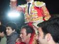 Un hombre en traje de torero sostiene un sombrero con una mano, mientras otros individuos lo rodean en una celebración nocturna.