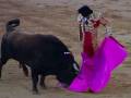 Un torero con un traje tradicional de color rojo y dorado, sostiene una muleta rosa mientras enfrenta a un toro negro en la arena de toros.