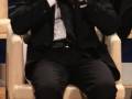 Un hombre en traje negro está sentado en una silla, con las manos cerca de su rostro. La imagen parece ser de un evento formal o conferencia, con otros asistentes en el fondo.