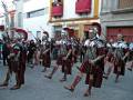 Desfile histórico con soldados romanos en traje tradicional, frente a edificios de estilo colonial.