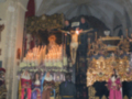 Un altar en una iglesia con estatuas religiosas y una cruz en el centro, rodeado de velas encendidas. Los personajes vestidos con trajes tradicionales se encuentran en el fondo, observando el altar.