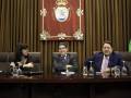 Tres personas sentadas en una mesa con un escudo real en el fondo.