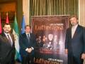 Dos hombres en trajes posan frente a un cartel de "Les Luthiers Lutherapia" con banderas y una imagen de un grupo musical en el fondo.