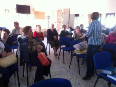 Una sala de conferencias con personas sentadas en sillas azules, una mujer hablando frente a un grupo. La pared tiene carteles y una lámpara.