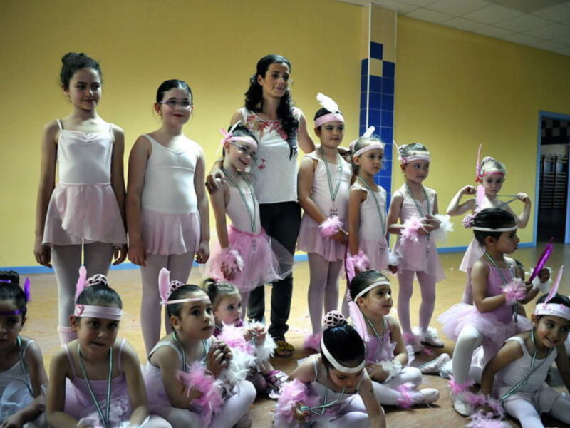 Grupo de niñas en trajes de ballet con plumas rosas, posando con una maestra en un estudio de danza.