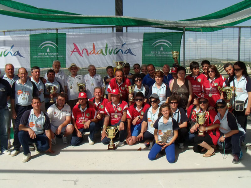 Fotografía de un grupo diversificado de personas posando juntos en una cancha deportiva, todos con trofeos y medallas. El fondo muestra un banner de Andalucía y un escudo con la palabra "Juegos". La imagen transmite una celebración deportiva.