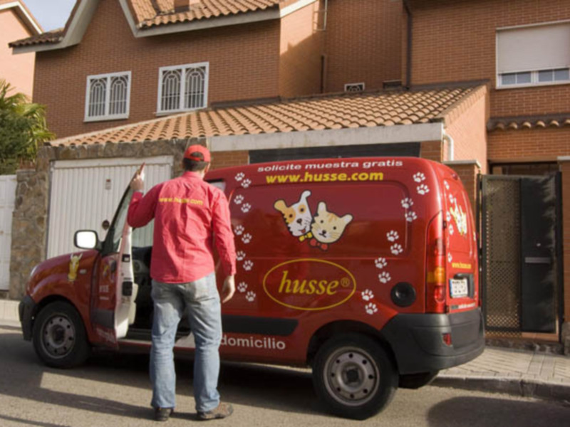 Un hombre con una chaqueta roja y un gorro de béisbol está abriendo la puerta de una camioneta roja con el logotipo de Husse. La camioneta está estacionada en una calle con casas de ladrillo rojo y un césped verde.