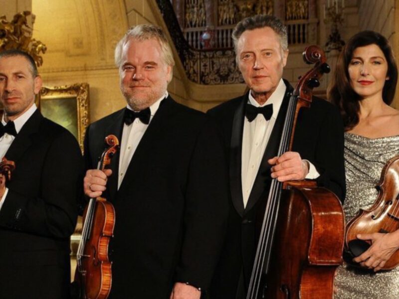 Una imagen de tres personas vestidas con trajes formales, posando junto a instrumentos musicales. La escena parece ser una representación de un grupo musical clásico, posiblemente para una presentación o concierto. La decoración en el fondo sugiere un entorno de lujo y elegancia, probablemente dentro de un palacio o sala de conciertos.