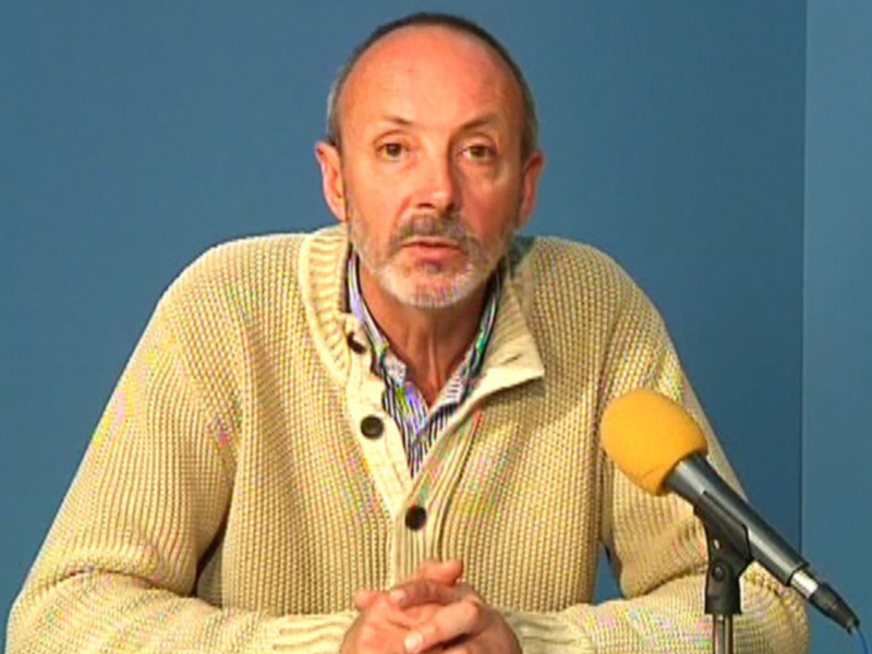 Persona hablando en un micrófono durante una presentación o entrevista en un entorno sencillo con pared azul.
