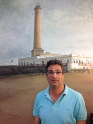 Se ve a un hombre con una camiseta azul posando frente a un faro alto y blanco. La escena se encuentra en una playa con arena, y el cielo está nublado.