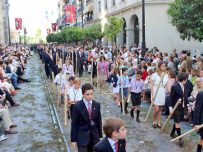 Una multitud de personas, incluyendo niños y adultos, participan en una procesión religiosa o festiva. Se observa a los participantes con sombrillas y vestimentas tradicionales, mientras caminan por una calle con árboles y edificios a ambos lados. La imagen captura la solemnidad y el fervor de la ceremonia.