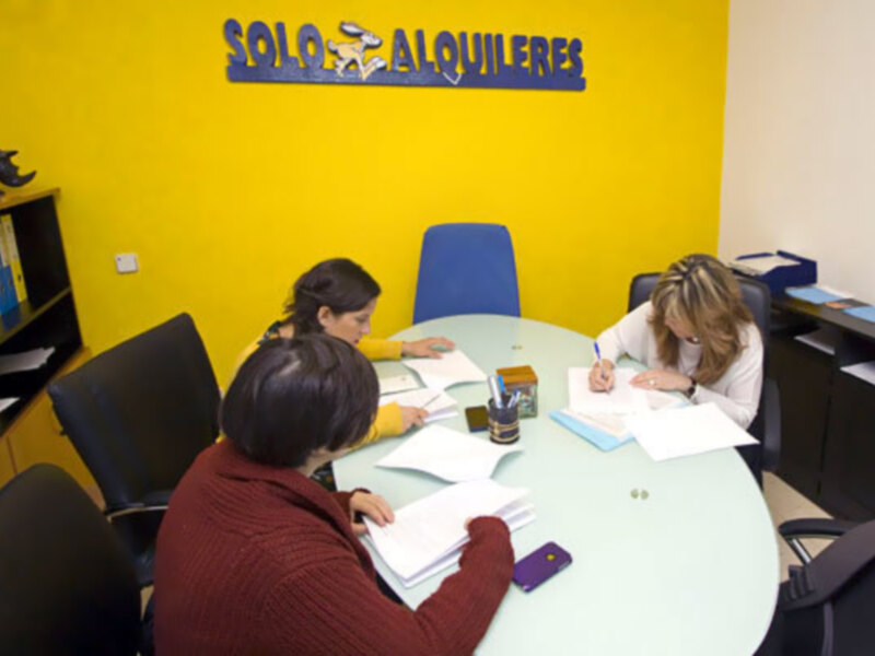 Tres personas trabajando en una oficina con una pared amarilla y un letrero que dice 'SOLO ALQUILERES'.