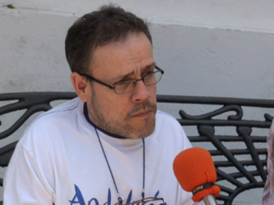 Un hombre con barba y gafas de lectura habla en un micrófono rojo. Se sienta en una silla metálica con fondo de pared blanca.