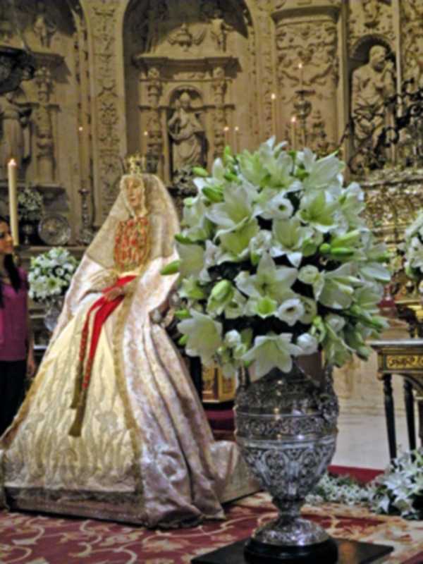 Una figura vestida con un manto dorado y una corona, posiblemente una imagen religiosa, se encuentra en un ambiente decorado con estatuas y flores. En el fondo, una estructura arquitectónica con detalles ornamentales y luces que iluminan el lugar.