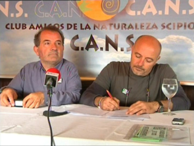 Dos hombres sentados en una mesa, uno escribiendo y el otro con un teléfono móvil. En la pared hay un cartel del Club Amigos de la Naturaleza.
