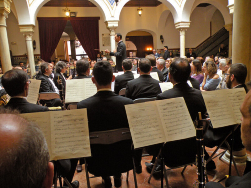 Una orquesta clásica en un auditorio con arcos y cortinas rojas. Los músicos están sentados, tocan instrumentos como clarinetes y violines, y tienen folletos de partitura en sus manos. La iluminación es cálida, y la atmósfera parece ser de una ceremonia o concierto formal.