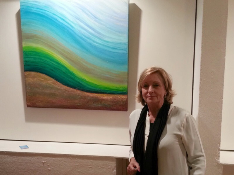 Una mujer con cabello corto y una blusa blanca posa junto a un cuadro abstracto de colores vibrantes. La imagen muestra una escena en un museo o galería, con la mujer sonriendo y el cuadro de fondo que parece una representación de un paisaje con tonos azules, verdes y marrones.