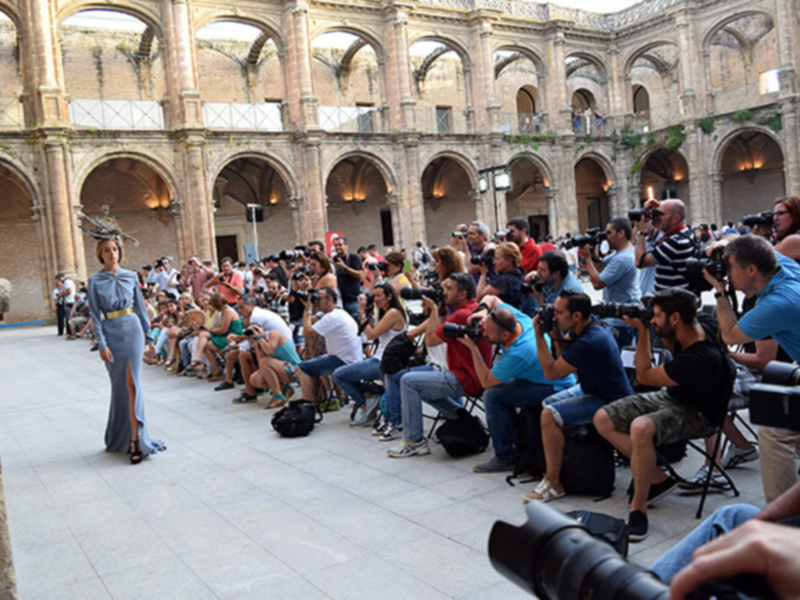 Desfile de moda en un patio histórico con una modelo en el centro y una multitud de fotógrafos alrededor.