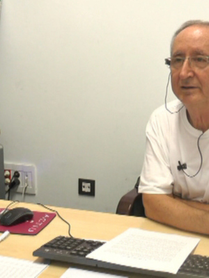 Un hombre con auriculares y una camiseta blanca está sentado frente a un ordenador en un escritorio. La pared detrás de él tiene una toma de corriente y un enchufe, con un estante de madera en la parte superior.