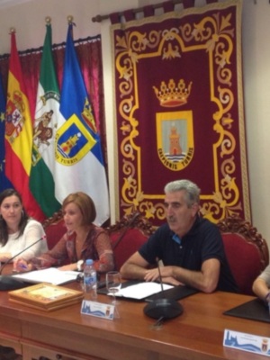 En una sala de conferencias, cinco personas se sientan en un escritorio frente a banderas y una gran bandera. La sala está decorada con plantas, cortinas rojas y un escudo heráldico en el fondo.