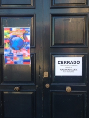 Puerta de madera oscura con dos paneles, uno de los cuales tiene un cartel informativo. Cartel muestra información sobre una actividad cultural y otro anuncia la apertura de la plaza Andalucía.