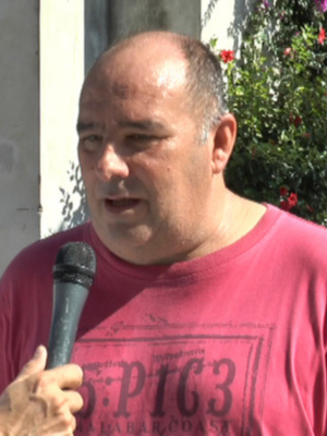 Un hombre con una camiseta roja está hablando en un micrófono. A su lado, hay una palmera y flores de color púrpura en el fondo.