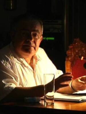 Un hombre en una oficina oscura, hablando en un micrófono. La iluminación es suave, enfocándose en su rostro y manos. En la mesa, hay documentos y un vaso de cristal vacío. La atmósfera sugiere una reunión formal o una conferencia de negocios.