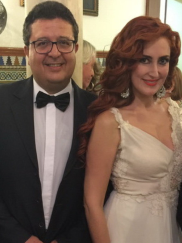 Una imagen de tres personas posando juntas en un ambiente festivo. La persona central es una mujer con cabello rojo, vestida de blanco y un hombre a su derecha también en traje formal. La pared al fondo tiene detalles de azulejos y una decoración tradicional.