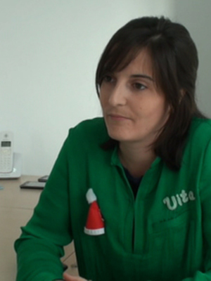 Una mujer en una oficina, con un suéter verde y una chaqueta roja con un logo. Está sentada frente a un escritorio con un teclado, una computadora y varios documentos en el fondo.