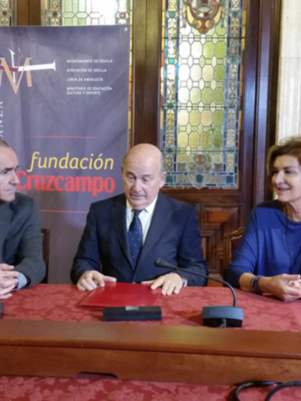 En una sala de conferencias con un fondo de vitrales y decoración clásica, tres personas se encuentran sentadas en una mesa de madera. La persona central lleva un traje oscuro y una corbata, mientras que los otros dos tienen vestimentas más informales. En el fondo, una pancarta de la Fundación Cruzcampo se puede leer claramente.