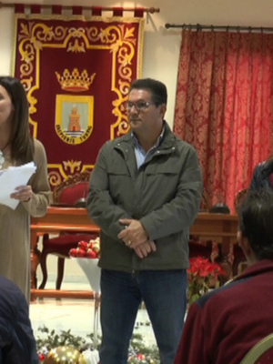 En una sala de conferencias, varias personas se encuentran en un evento formal. La imagen muestra a dos mujeres y un hombre hablando frente a una audiencia de personas sentadas en sillas. La sala está decorada con banderas y un escudo heráldico, indicando una celebración o reunión oficial. La atmósfera es formal y el ambiente parece ser de una institución o organización importante.
