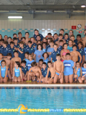 Grup de natació infantil a una piscina. Estan posant per a una foto grup al costat d'una piscina. Els nens i nenes estan vestits amb uniformes de natació. Al fons, hi ha una parella d'escorxadors i un calendari amb la data.